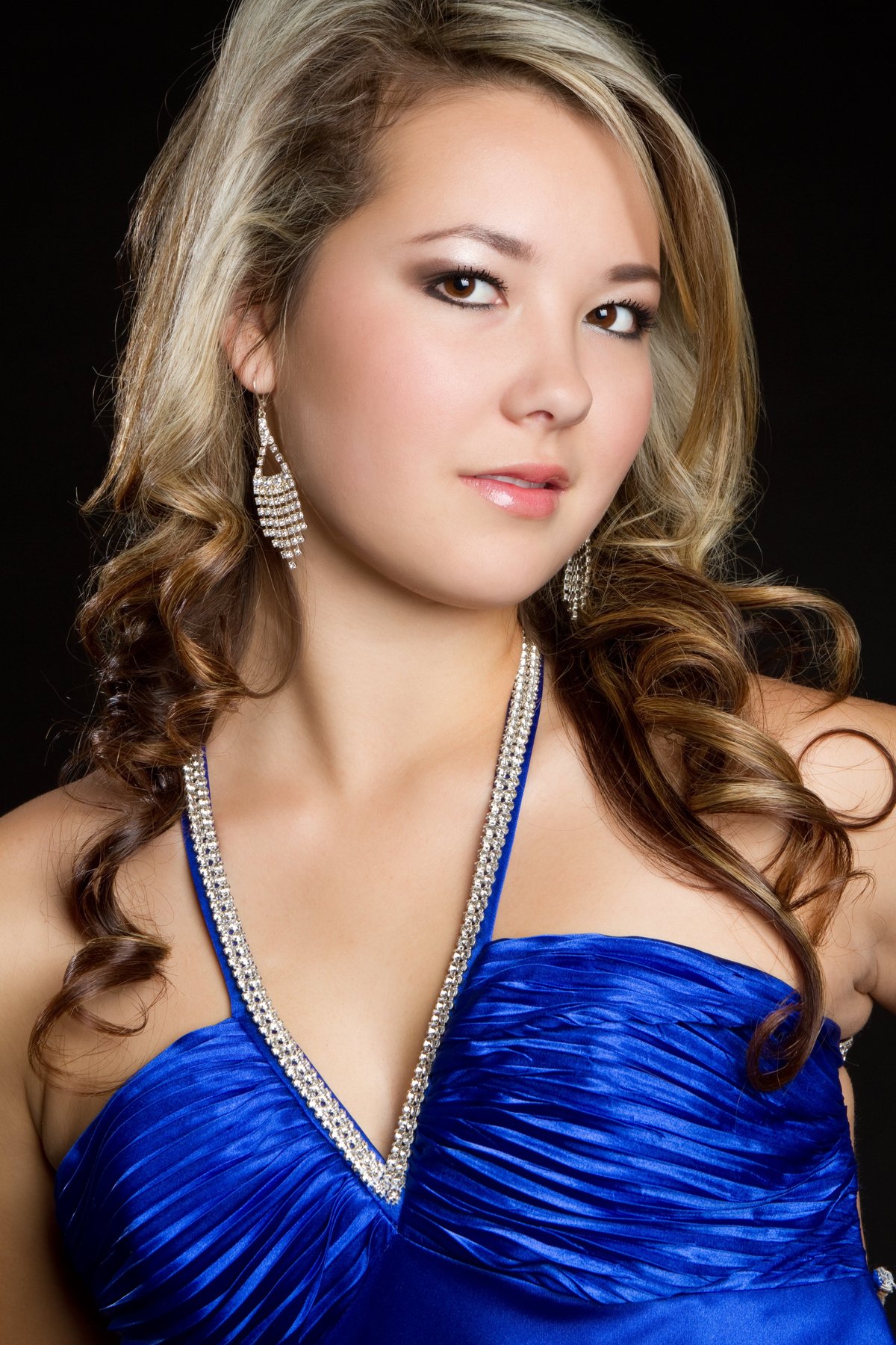 Woman in blue formal dress with crystal necklace and earrings, looking over shoulder against black background