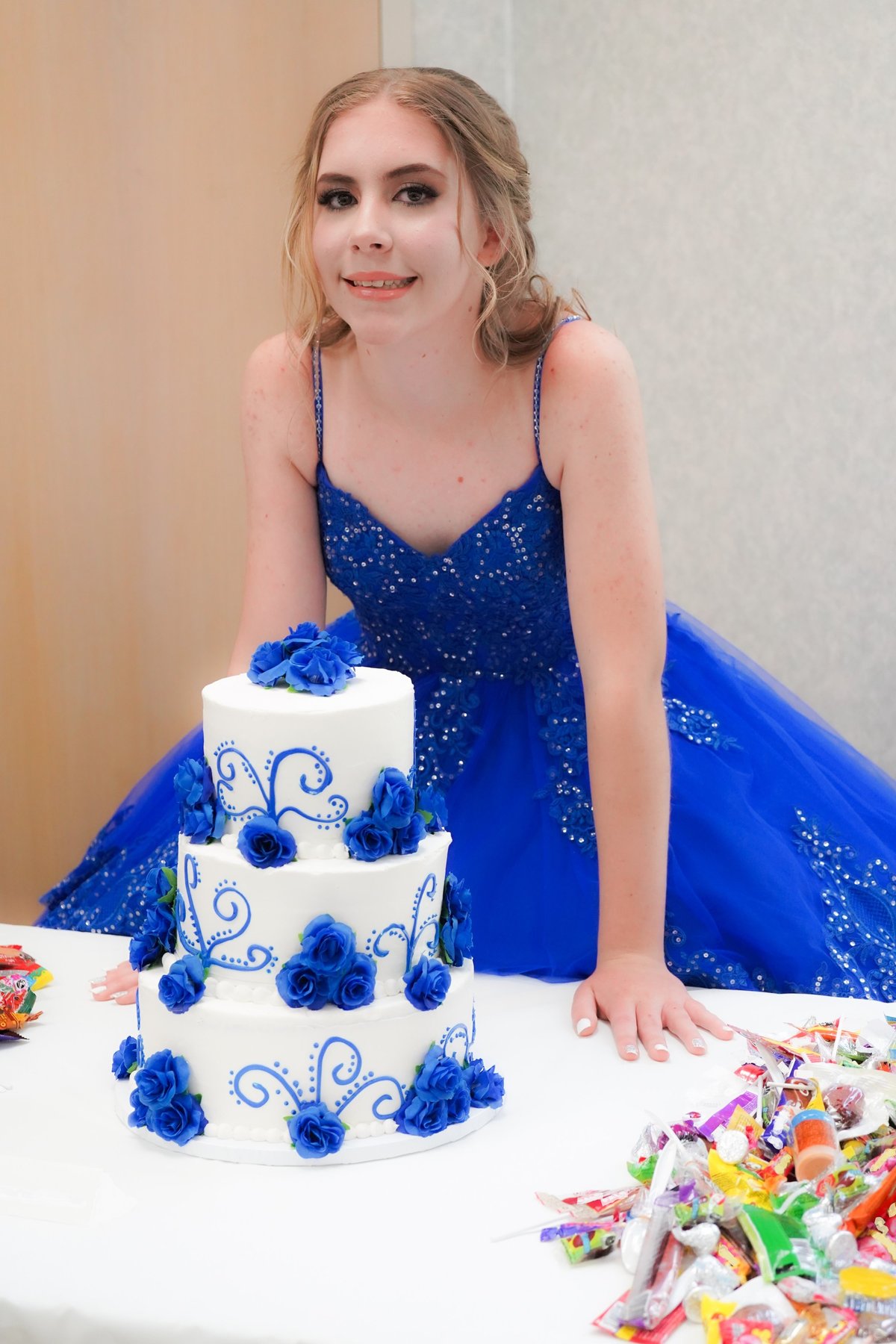 A teen girl, wearing a royal blue ball gown for her quinceañera, smiles in front of three-tiered birthday cake.