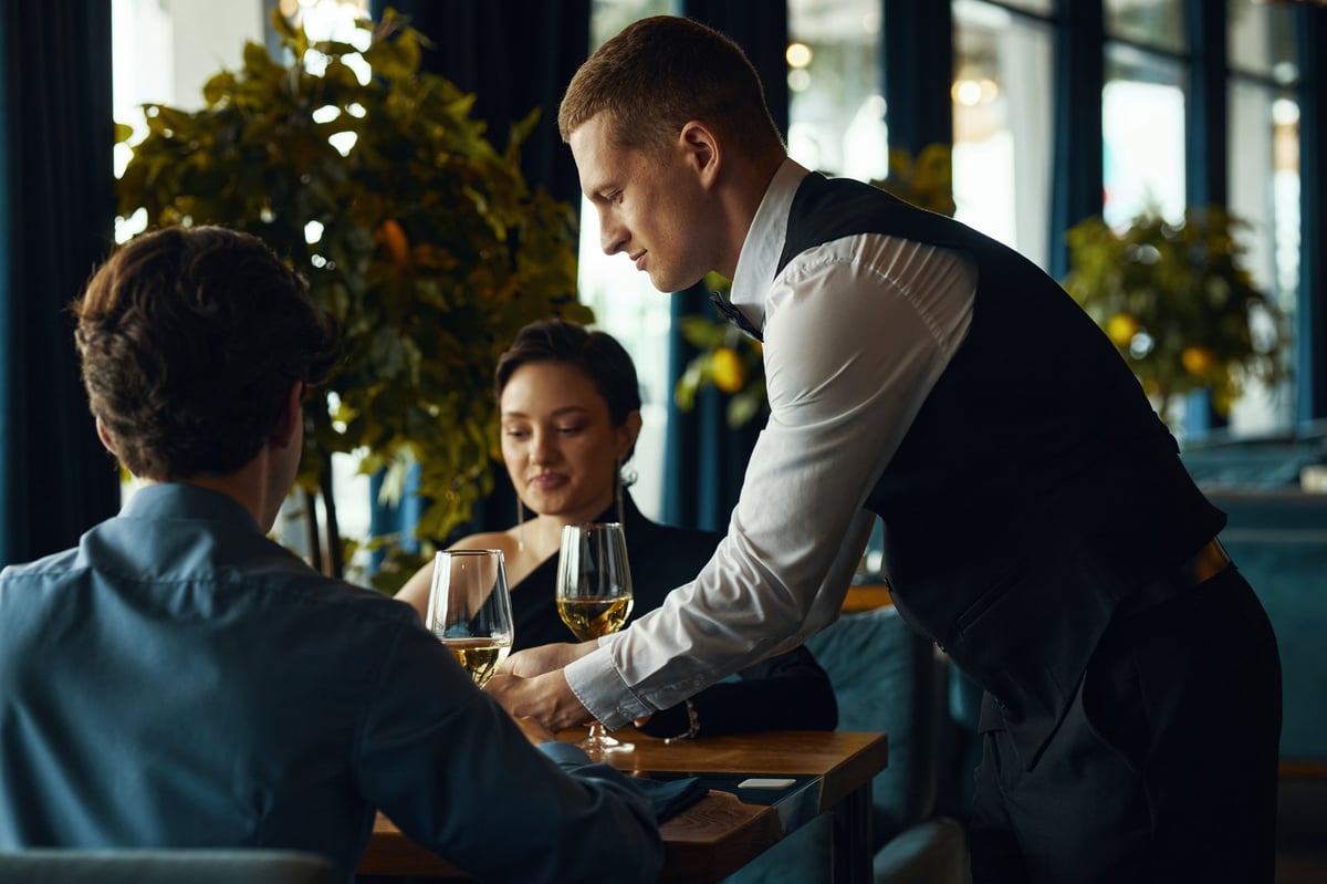 Young adult man serving wine to a couple sitting at restaurant table