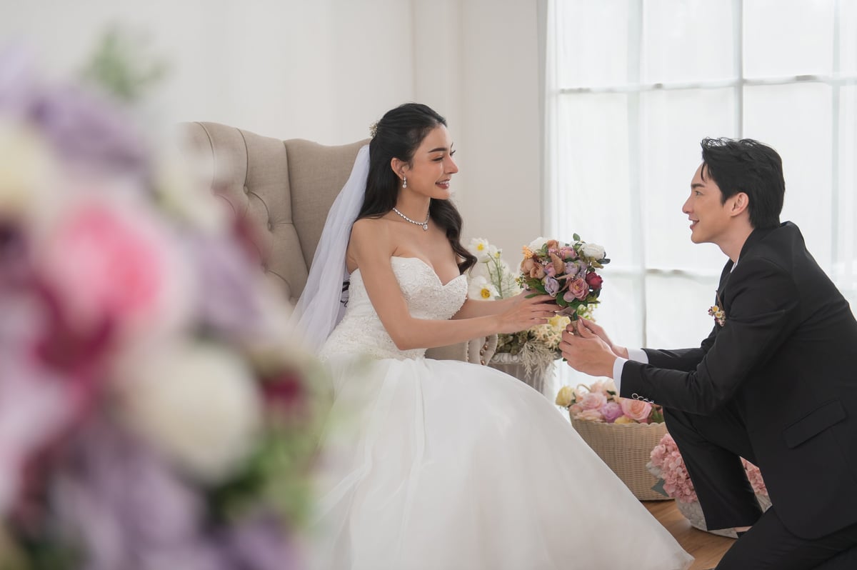 Groom kneels and offers a bouquet to his smiling bride in a white wedding dress, surrounded by floral decorations