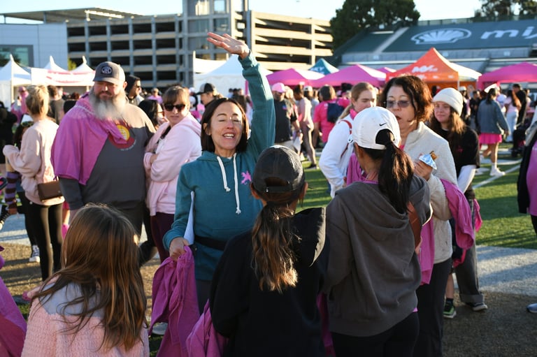 Large crowd of participants wearing pink and purple at the Sacramento Breast Cancer Walk hosted by Kaiser Permanente, on a grassy field with tents and urban buildings in background